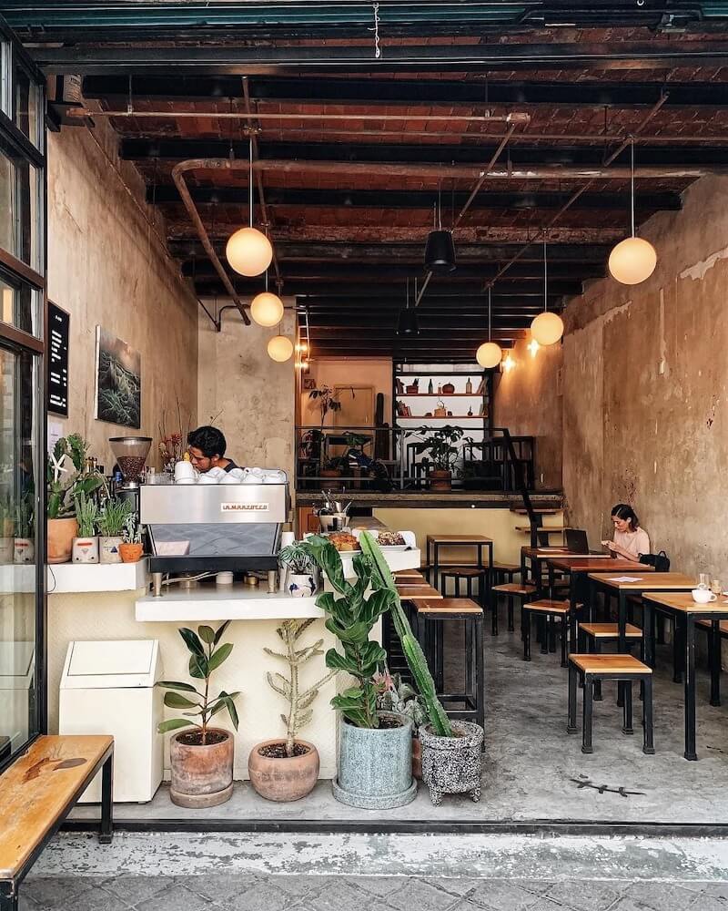 Vẻ đẹp thô mộc của những bức tường loang lổ và trần gạch cũ hòa quyện tinh tế với ánh đèn vàng ấm áp và sức sống của cây xanh, tạo nên một không gian cafe đầy chất thơ và gần gũi
