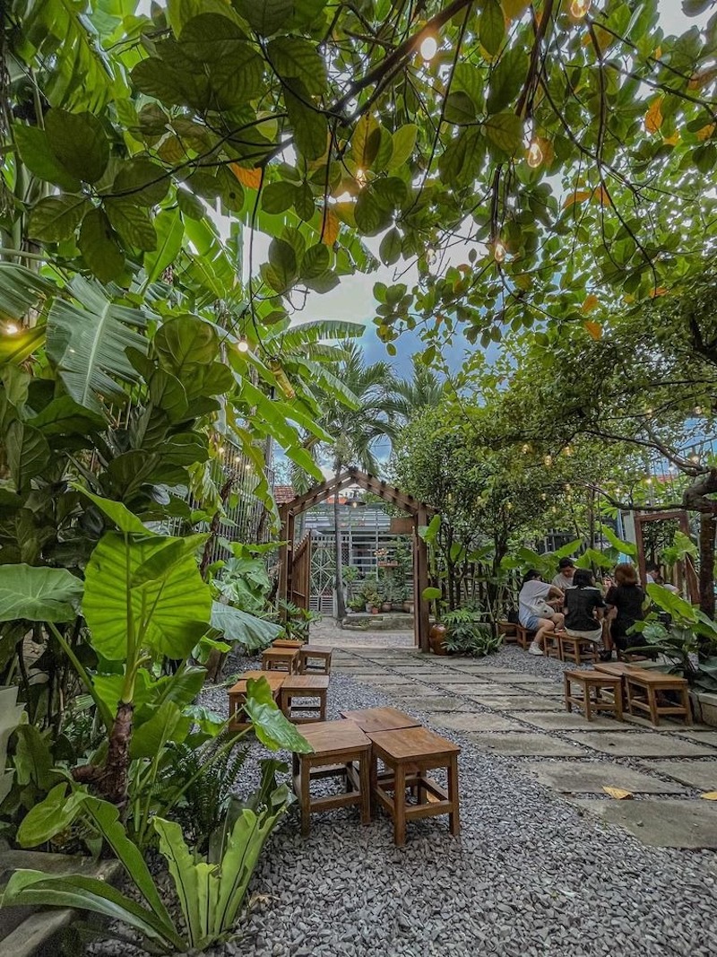 ’Thiết kế quán cafe bình dân kiểu sân vườn nhỏ tận dụng tối đa cây xanh, lối đi lát gạch giản dị và bàn ghế gỗ mộc mạc để tạo cảm giác gần gũi, thoải mái