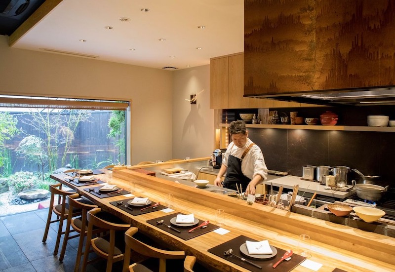 Quầy bếp mở kết hợp bàn ăn dài bằng gỗ, cho phép thực khách quan sát trực tiếp quá trình chế biến trong khung cảnh yên tĩnh và tinh tế 