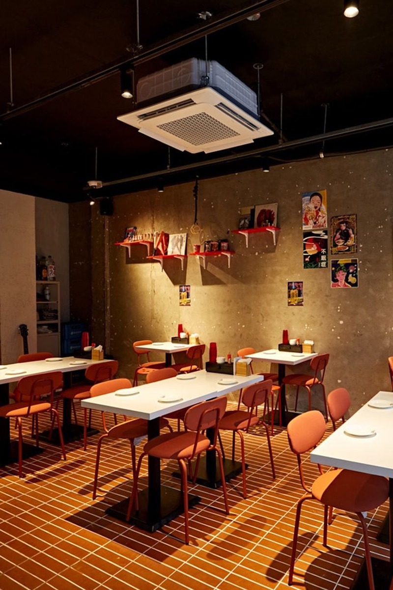 Bàn ghế tông cam nổi bật trên nền gạch đỏ, kết hợp tường bê tông trần và tranh treo tạo cảm giác trẻ trung, cá tính 
