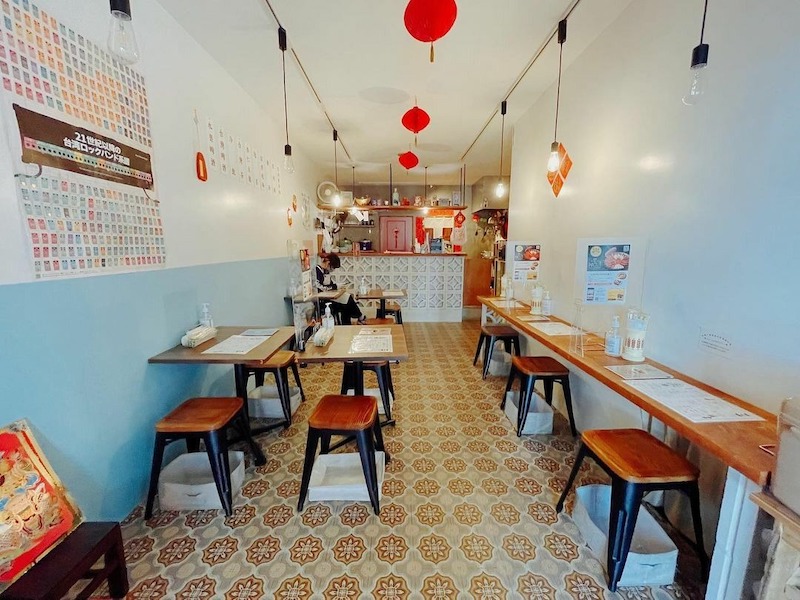 Bố cục dài gọn với bàn gỗ áp tường, ghế đôn thấp, sàn gạch bông hoài cổ và đèn lồng đỏ dẫn mắt thẳng về quầy phục vụ phía cuối 