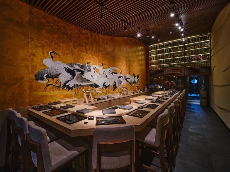 Mẫu nhà hàng Omakase sang trọng với quầy bar gỗ hình chữ U, mảng tường nghệ thuật và hệ trần nan gỗ tinh tế dưới ánh đèn spotlight ấm cúng