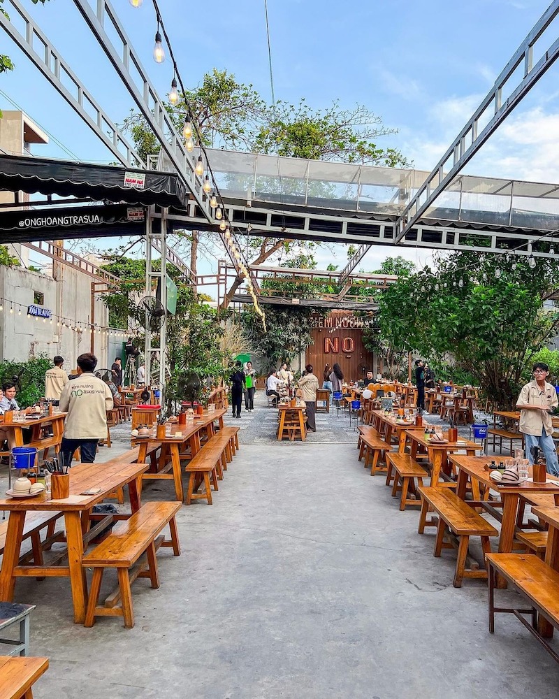 Không gian mở, bàn ghế gỗ dài và lối đi rộng rãi, kết hợp cây xanh và đèn dây tạo cảm giác thoáng mát, gần gũi và dễ chịu cho thực khách