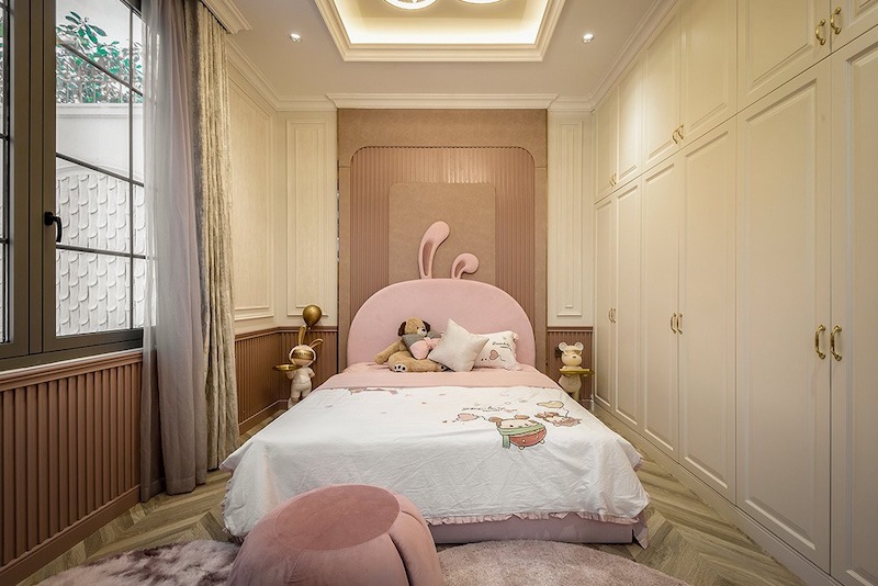Giường bọc nỉ hồng dáng cong kết hợp hệ tủ âm phào chỉ tinh tế, ánh sáng trần dịu và vật liệu êm ái tạo cảm giác ấm áp, an toàn, đáng yêu nhưng vẫn sang