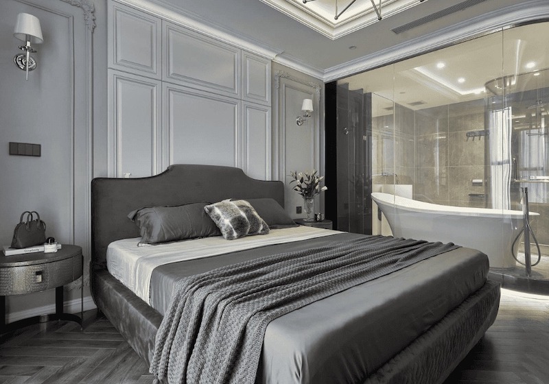 Giường bọc nỉ xám dáng cong mềm, vách tường phào chỉ cổ điển tinh gọn, tab đầu giường kim loại và không gian kính sau giường tạo cảm giác sang trọng, hiện đại