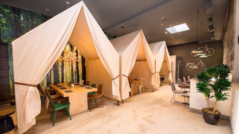 Không gian quán thiết kế như khu camping trong nhà, với các lều vải trắng, bàn gỗ mộc, ánh sáng ấm, tạo cảm giác trải nghiệm gần gũi thiên nhiên