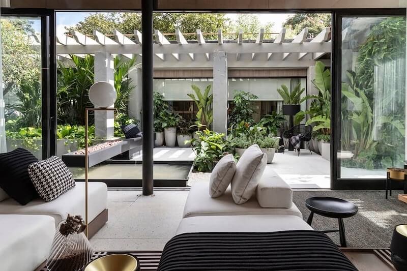 Không gian phòng khách mở hoàn toàn ra sân trong xanh mát, cây cối bao quanh, ánh sáng tự nhiên tràn ngập, thể hiện rõ tinh thần biophilic hiện đại, thư giãn và kết nối thiên nhiên