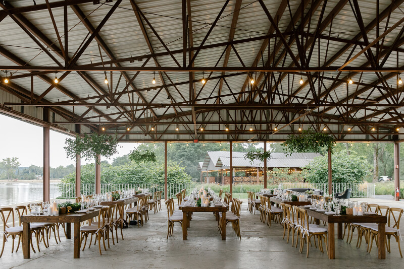 Không gian sảnh tiệc ngoài trời sử dụng kết cấu khung thép mái cao, bố trí bàn dài mộc mạc hướng ra cảnh quan tự nhiên, tạo cảm giác thoáng đãng và gần gũi