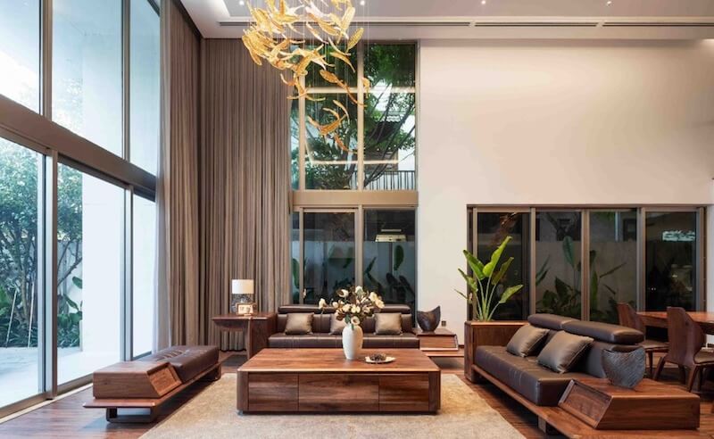 Trần cao thông tầng và mảng kính lớn ngập sáng, nổi bật với bộ sofa gỗ óc chó dáng thấp, đường nét vuông vức kết hợp vân gỗ nâu trầm sang trọng tạo cảm giác bề thế 