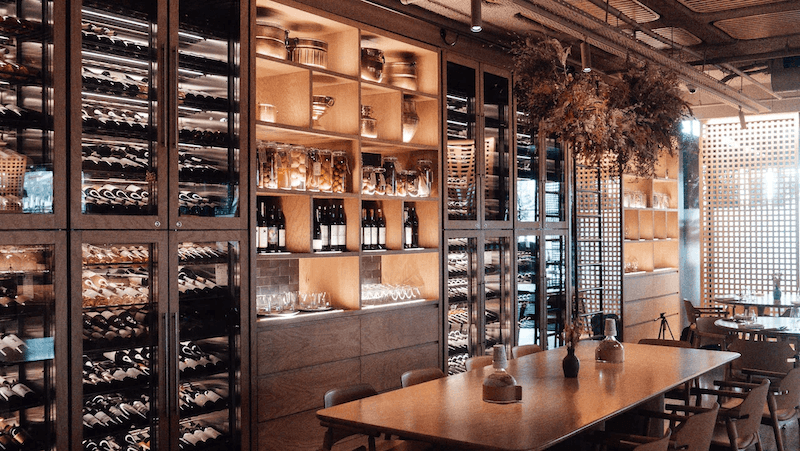 Không gian wine lounge hiện đại với tủ rượu kính kịch trần, ánh sáng ấm, bàn gỗ dài tạo trải nghiệm sang trọng, tinh tế và gần gũi