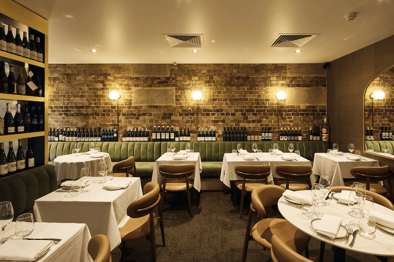 Không gian nhà hàng rượu vang ấm cúng với tường gạch trần, ánh sáng vàng dịu, bàn phủ khăn trắng và ghế da tạo cảm giác sang trọng, tinh tế
