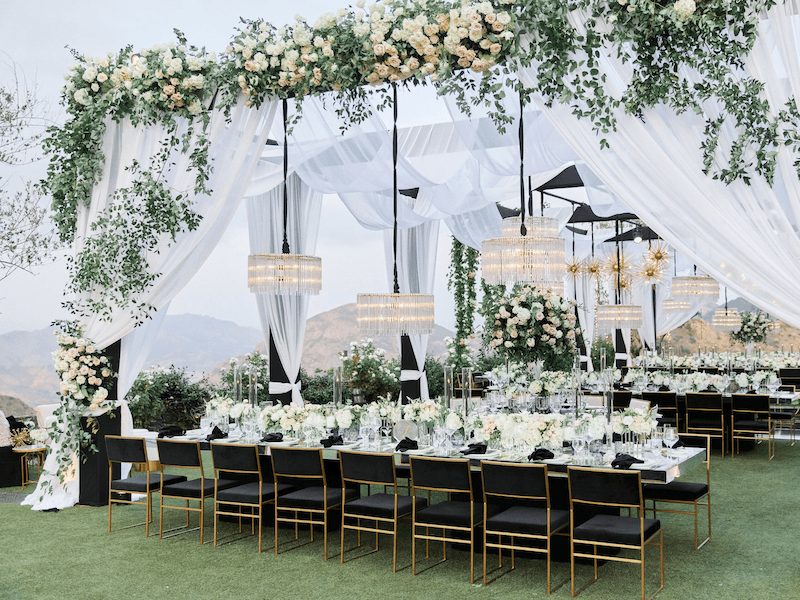 Không gian Garden Wedding nổi bật với mái vòm hoa phủ xanh, rèm voan trắng bay nhẹ, đèn chùm pha lê treo giữa thiên nhiên và bàn tiệc sang trọng giữa khung cảnh núi đồi