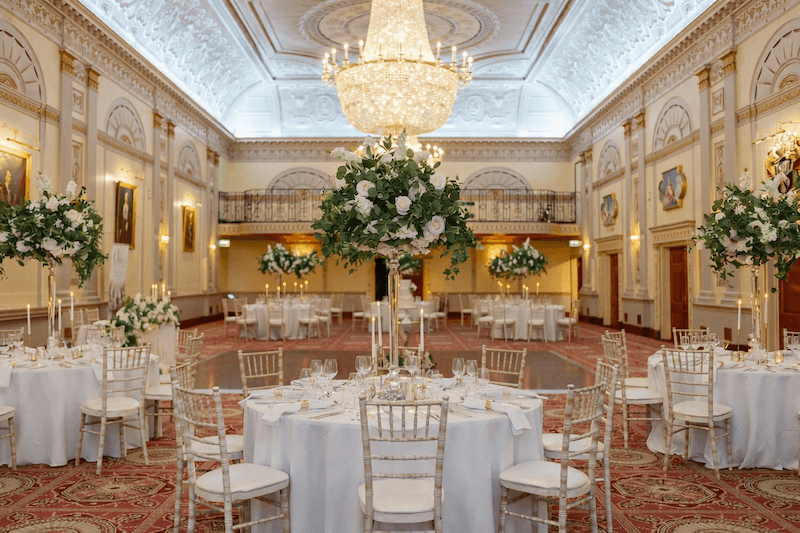 Sảnh tiệc Tân cổ điển nổi bật với trần hoa văn chạm nổi tinh xảo, đèn chùm pha lê trung tâm, cột đối xứng thanh lịch và bàn tiệc trắng sang trọng