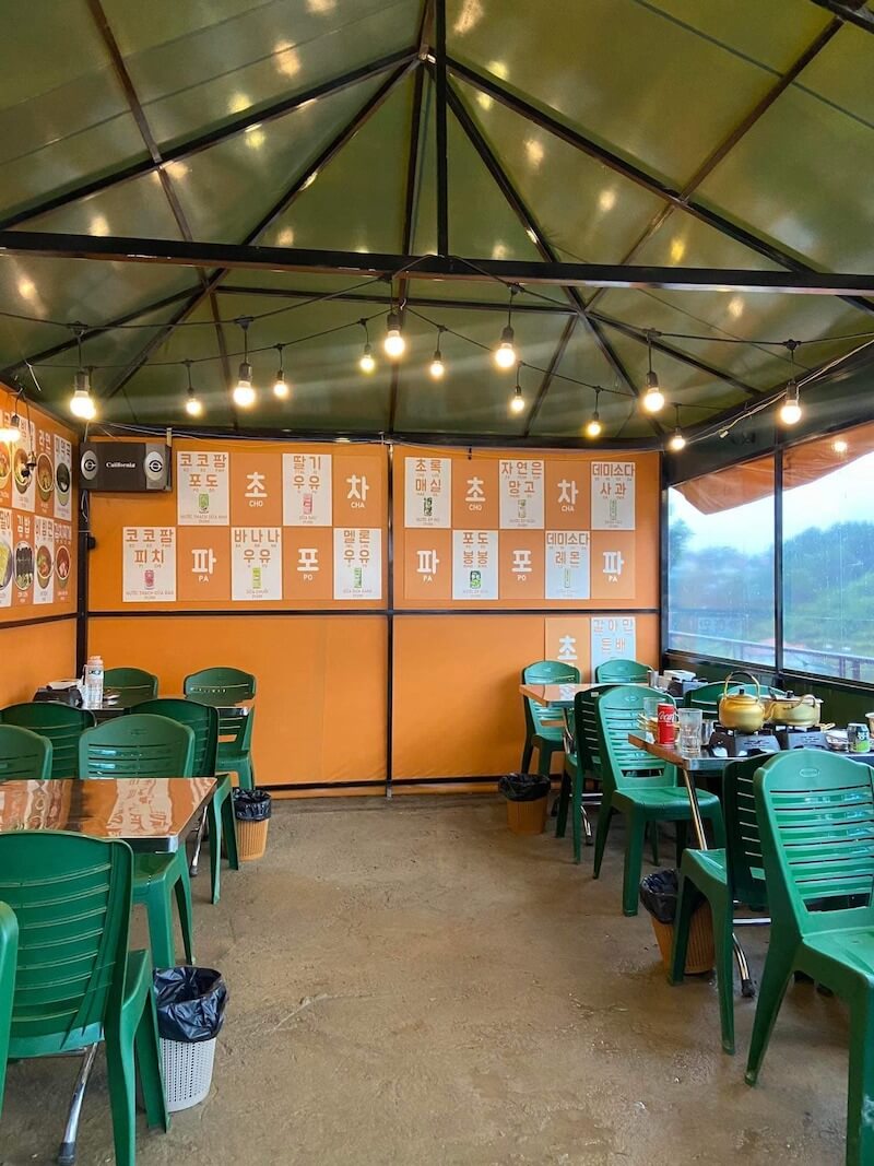 Mảng tường cam nổi bật phủ kín poster menu tiếng Hàn mang đậm chất đường phố, đóng vai trò vừa là bảng hiệu vừa là background check-in đúng chất K-culture