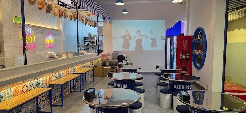 Hệ bàn thùng phi kim loại in chữ “POCHA” kết hợp ghế đẩu tròn thấp, mô phỏng đúng tinh thần quán nhậu vỉa hè Hàn Quốc nhưng được “nâng cấp” sạch sẽ và gọn gàng hơn