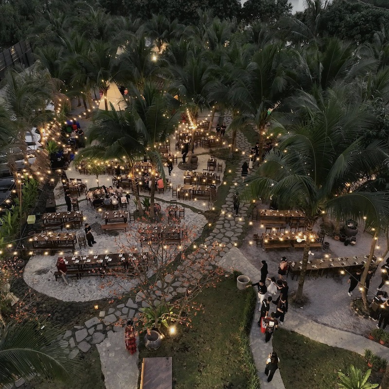 Sàn gỗ ngoài trời tông nâu ấm kết hợp bàn ghế gỗ khung sắt đơn giản, bố trí linh hoạt giữa các thân cây lớn tạo cảm giác gần gũi thiên nhiên