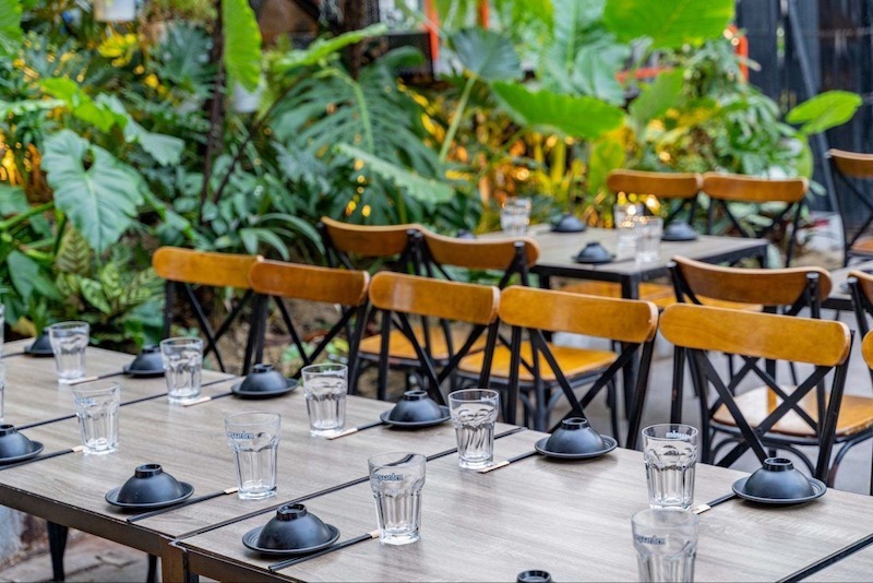 Quán sân vườn mang đậm tinh thần tropical với mật độ cây xanh dày, tán cây lớn tạo bóng mát tự nhiên, kết hợp lối đi uốn lượn bằng sỏi