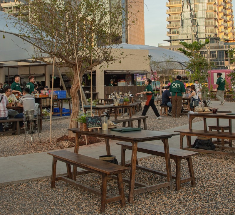 Không gian quán sân vườn phong cách tropical, bàn gỗ sáng màu, cây xanh tán lớn, ánh đèn treo ấm áp, bố cục mở, mang cảm giác thư giãn