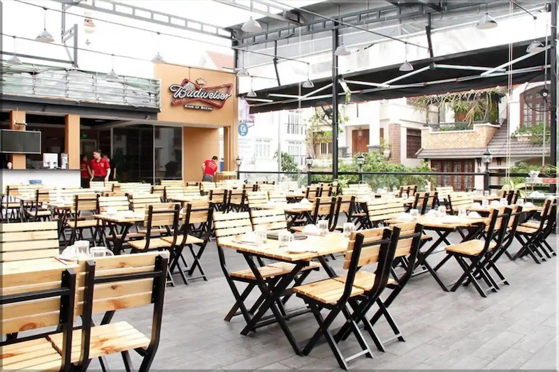 Quán nhậu sân vườn nhiều cây xanh lớn, bàn ghế sắt gọn nhẹ, ánh sáng vàng buổi tối tạo không khí mát mẻ, thư giãn, ngồi lâu