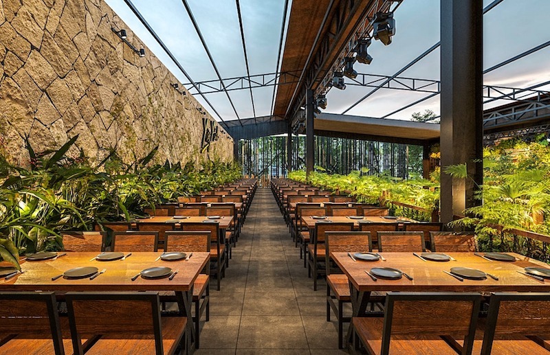 Bố cục bàn ghế thẳng hàng tạo trục nhìn rõ ràng, kết hợp mái che thép nhẹ và mảng xanh dày hai bên mang lại cảm giác thoáng, mộc và chỉn chu