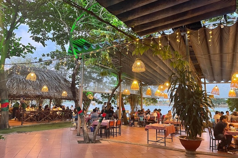 Hệ mái che nhẹ kết hợp đèn tre thả ấm áp, xen kẽ cây xanh và bàn ghế gỗ mộc tạo cảm giác gần gũi, thư giãn, phù hợp tụ họp buổi