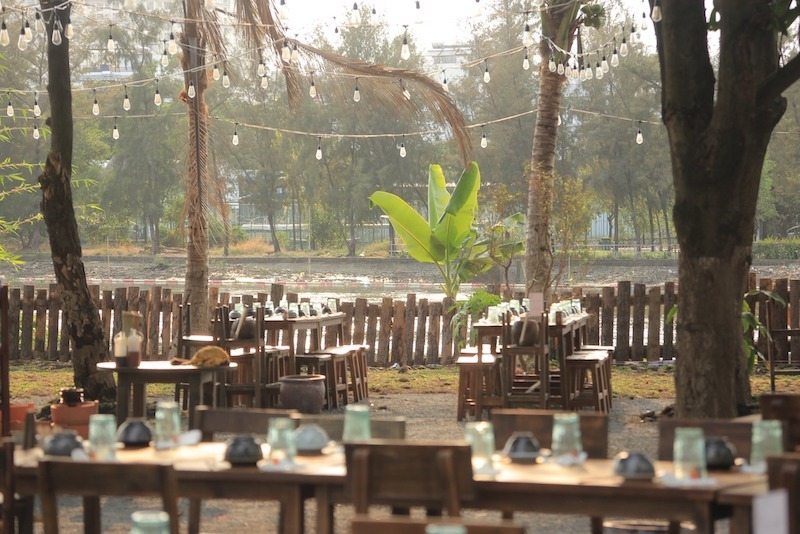 Bàn ghế gỗ mộc bố trí dưới tán cây, đèn dây treo cao tạo ánh sáng dịu, mở tầm nhìn ra mặt nước, mang lại cảm giác thư giãn, gần gũi thiên nhiên